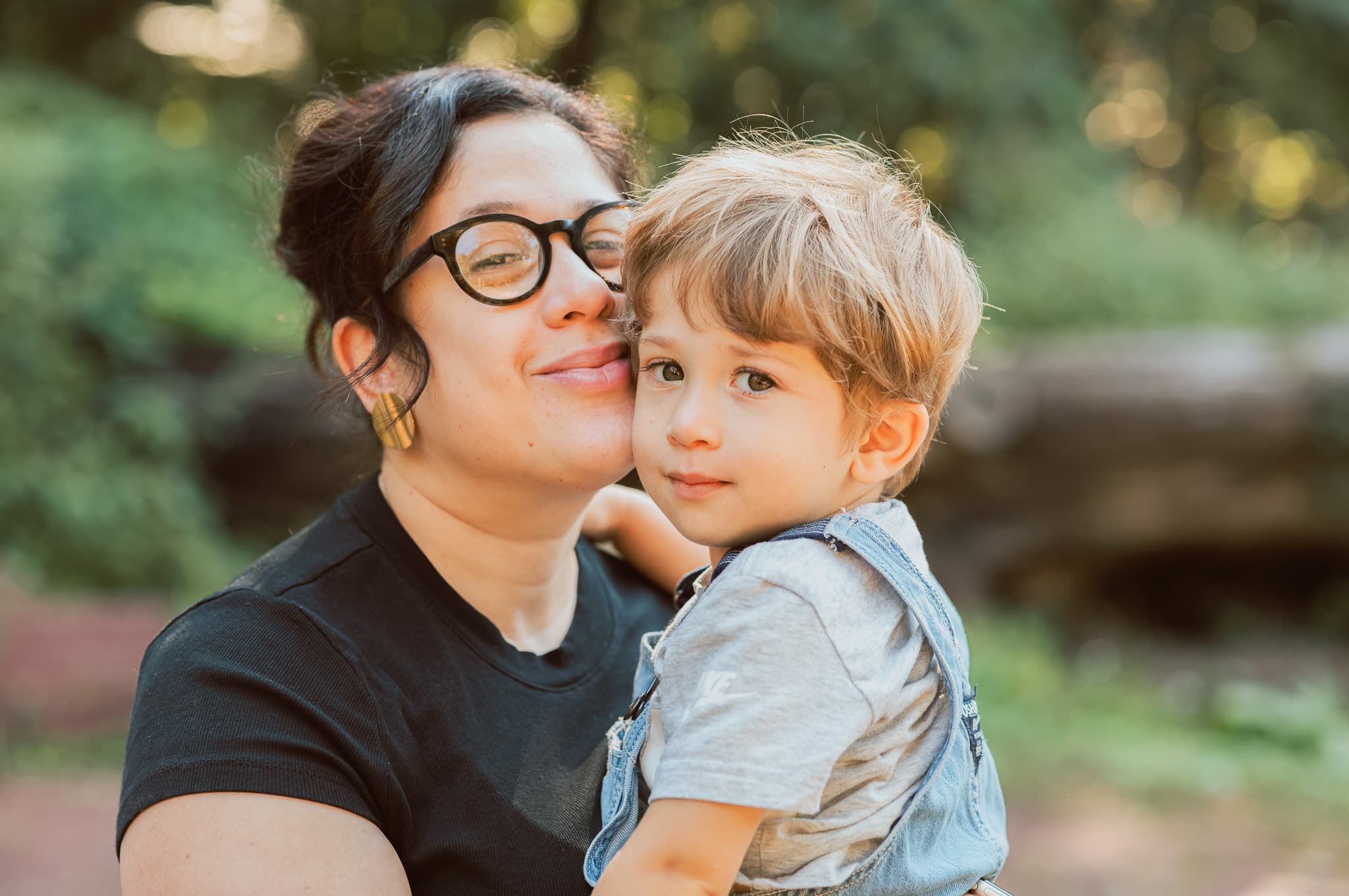Mother in glasses holding her young son cheek to cheek, outdoor family portrait in a wooded park by Fuentes Studio