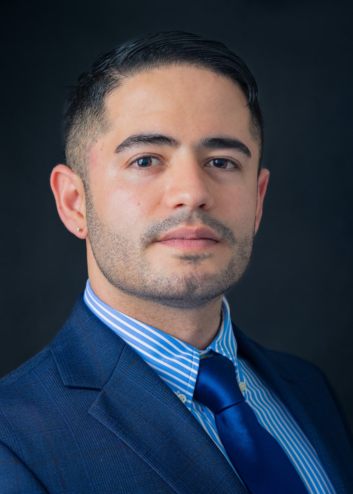 Man with a beard in a navy suit, striped shirt and blue tie, corporate headshot on a dark backdrop